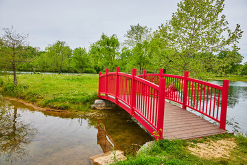 Gorgeous fire engine red walking bridge onto green island over stream leading to pond and geese