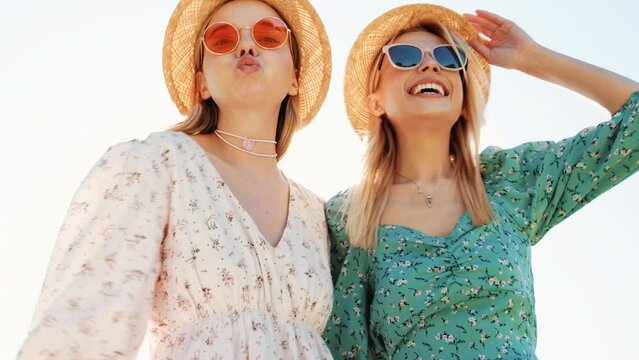 Two young beautiful smiling hipster woman in trendy summer sundress. Carefree women posing in street in hats. Positive models having fun and hugging at sunset outdoors. Cheerful and happy. Bottom view