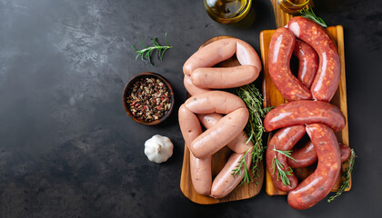 Assorted fresh sausages with thyme, rosemary, olive oil and garlic on a wooden chopping Board on a dark concrete table. Ready to cook sausages for dinner on black background. Top view with copy space