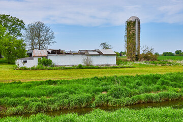 Farmland with runoff ditch summer corn grain silo covered in ivy and roof caved in of building