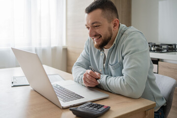 Portrait of young smiling man using laptop sitting at desk, writing in notebook. Cheerful guy browsing internet, watching webinar studying online, looking at pc screen at home.