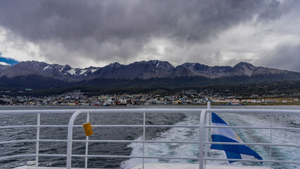 Behind the stern of the tourist boat there is a foam trail. The blue - white flag of Argentina is flying on the railing. The buildings of Ushuaia are visible on the shore. A mountain range, cloudy sky
