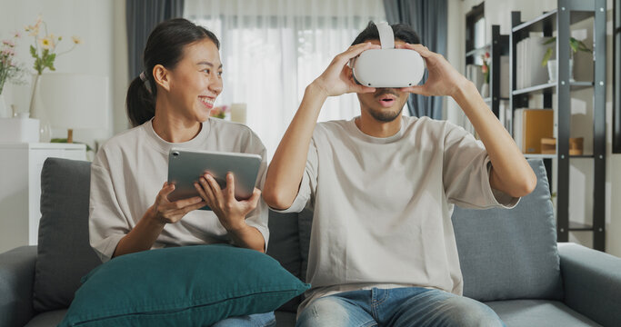 Young Asian couple sit on couch hold digital tablet and wear virtual reality glasses are watching and show imagine spend time together have fun at home on weekend. Lifestyle tech innovation concept.