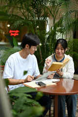 Two Asian students are sitting at a cafe in an outdoor area, preparing for the exam together