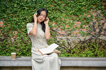 A happy Asian woman enjoys her music on her headphones while relaxing on a bench