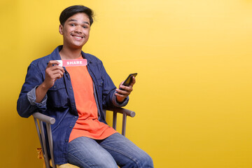 Young content creator male holding cell phone while showing printed paper with SHARE text, asking...