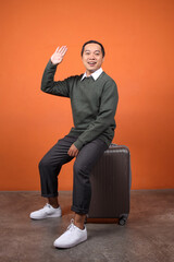 Full length of Asian man waving hand saying hi goodbye while sitting on luggage ready to go on vacation isolated over orange background © Gatot