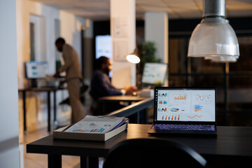 Laptop computer with financial data graphs standing on desk table in startup office, coworkers working late at night at company strategy. African american employees brainstorming business ideas