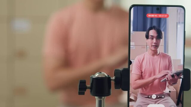 Close up youth Asia male owner small business online shop sit front of phone look at camera live streaming see comment on digital tablet from customer sales promotion at retail online shop at home.