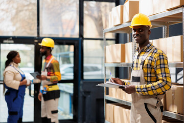 Shipment service warehouse manager checking orders list on clipboard portrait. Smiling young african american delivery operator working in storehouse and looking at camera