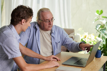 Senior man asking grandson to help him with installing applications on laptop - obrazy, fototapety, plakaty