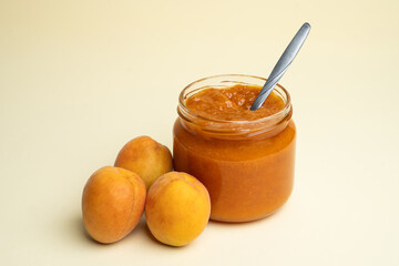 Apricot jam in a glass jar with a spoon inside, fresh apricots on a yellow background