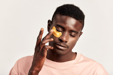 Close up portrait of young handsome black guy applying cosmetic under eye hydrogel patch on his...