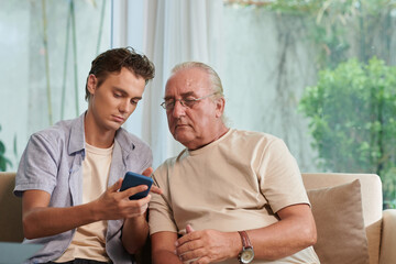 Young man showing senior man how to send text messages, voice messages and stickers in messenger