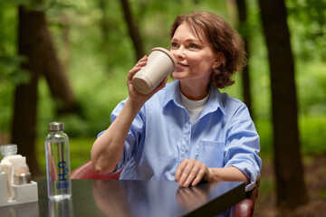 Obraz premium Mature woman sitting in cafe at the table and drinking coffee from a paper cup. Senior lady enjoying life on the summer terrace. Attractive middle-aged brunette in a blue shirt with a dreamy smile.