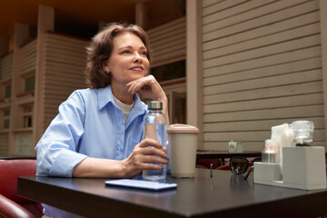 Attractive mature woman sitting in cafe at the table with bottle of water and paper cup of coffee. Senior lady enjoying life on the summer terrace. Middle-aged brunette in blue shirt smiling dreamily.