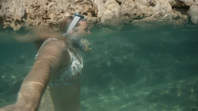 The camera captures two perspectives simultaneously, showing both above and below the sea surface. A young woman wearing a snorkeling mask and a breathing tube can be seen, with underwater rocky cliff