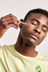 Young handsome black guy applying beauty serum on his face. African American millennial man uses beauty products maintaining his natural beauty with skincare routine. Men's self-care.