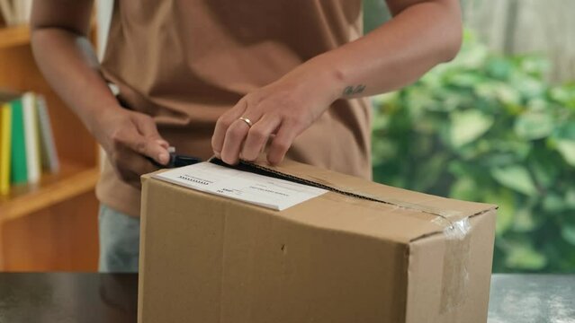 Close-up of woman unpacking box parcel with cutter knife at home