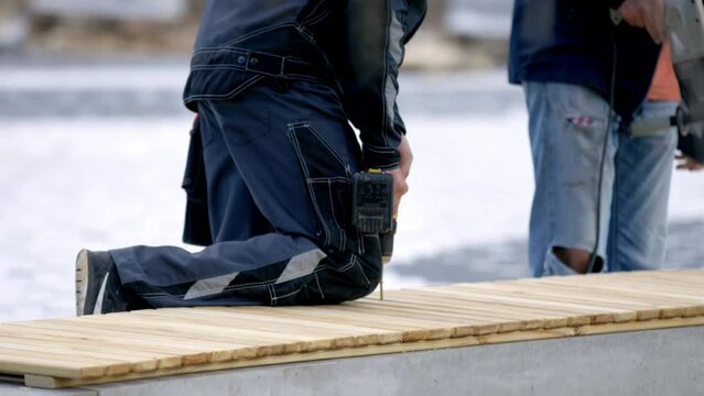 Workers use drill to make holes in bench wooden boards at daytime. Two employees secures boards of wood together to construct a bench during renovation of park