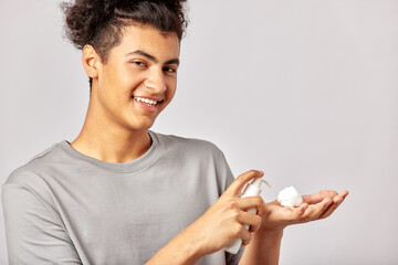 Happy young smiling guy putting facial wash on his fingers, preparing to apply the beauty product on his face. Swarthy man with black curly hair uses hydrating skin cleanser in his skincare routine.