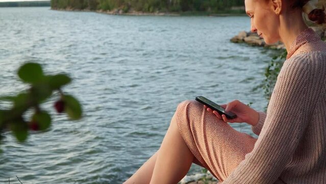A girl in a dress sits on the shore of the lake. A young woman is using her phone. She takes pictures and sends messages over the Internet via her smartphone.