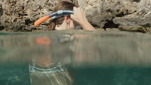 A young woman is in the sea among the rocks. She puts on her mask and dives underwater. The camera follows her