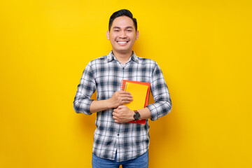 Smiling happy handsome young Asian man wearing a white checkered shirt holding books on hand isolated over yellow background. People Lifestyle Concept