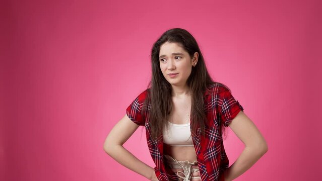 Distressfully looking casually dressed young woman with long dark hair touching her tummy with both hands apparently experiencing acute stomach pain against pink background