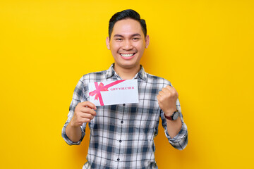 Excited young Asian man wearing a white checkered shirt holding a gift voucher and celebrating the winner isolated on yellow background. People Lifestyle Concept