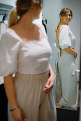 Fototapeta premium Adorable woman in new white blouse and being trousers smiling while checking outfit in reflection in dressing room.