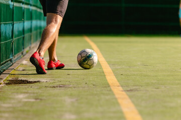 A soccer player in red sneakers kicking a ball on a green artificial football field behind a yellow...