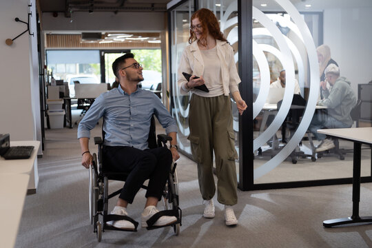 Young business colleagues, collaborative business colleagues, including a person in a wheelchair, walk past a modern glass office corridor, illustrating diversity, teamwork and empowerment in the