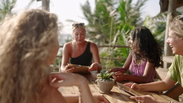 Diverse female friends in outdoor restaurant sipping drinks reading menu. Multiracial cheerful girls at the table in tropical cafe chatting deciding on food. Young multiethnic women having dinner