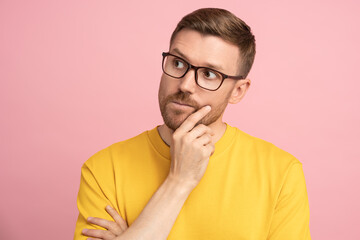 Thoughtful puzzled American man look away holding hand on chin. Pensive student or entrepreneur in eyeglasses ponder making decision of investment looking at empty copy space over studio pink wall