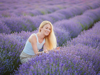 Lavender field. Young attractive woman with blond hair walking between rows of lavender flowers, posing for camera, relaxing, resting, enjoying nature, smelling and touching flowers. Mental health