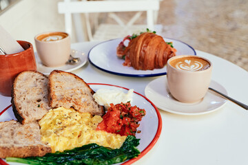 Brunch in an outdoor cafe - bread, scrambled eggs, tomatoes, butter, coffee on a white table