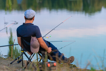 A young Caucasian male fisherman sits on a chair on the shore and catches fish. View from the back. No face. Evening or morning outdoors.