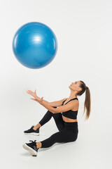 Fototapeta na wymiar woman in black sporty outfit throwing fitness ball and sitting on white background, athletic concept