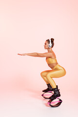Fototapeta na wymiar balance, woman in kangoo jumping shoes and wireless headphones exercising on pink background, squats