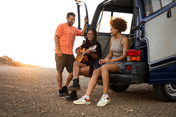 A group of 3 young multetnicos are sitting in the back of a camper van having fun with a flamenco guitar at sunset. Concept of camping with a camper van. Traveling with multiracial friends.Diversity.