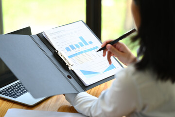 Over shoulder view of female economist analyzing sales statistics document, using laptop at her office desk