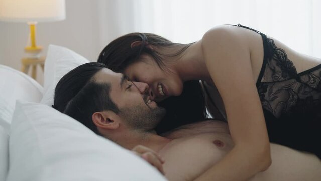 Romantic sexy moments lifestyle for a young couple at home, hot Asian lady wearing black bra teasing young aroused man on bed, passionate couple enjoying sensual foreplay petting before making love