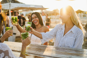 Happy girls cheering with mojitos on the beach at chiringuito sunset party - Summer vacation and...