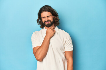 Long-haired man in a white polo, blue studio suffers pain in throat due a virus or infection.