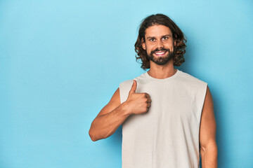 Bearded man in a tank top, blue backdrop smiling and raising thumb up