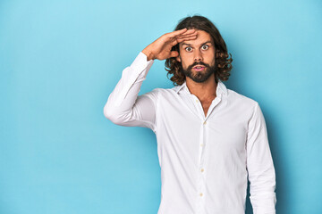 Bearded man in a white shirt, blue backdrop shouts loud, keeps eyes opened and hands tense.