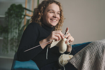 Curly haired woman sitting on the sofa in the living room and knitting a scarf for the winter....