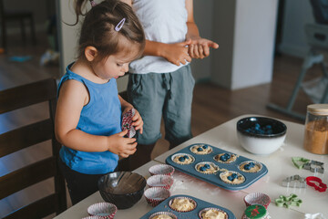Two children learning to cook, pouring the dough into special molds for baking portioned cupcakes,...