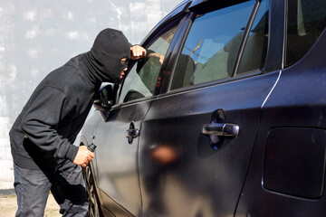 Man dressed in black wearing mask hold screwdriver, looking through car window and wondering how to break into the car. Car thief, car theft concept - obrazy, fototapety, plakaty